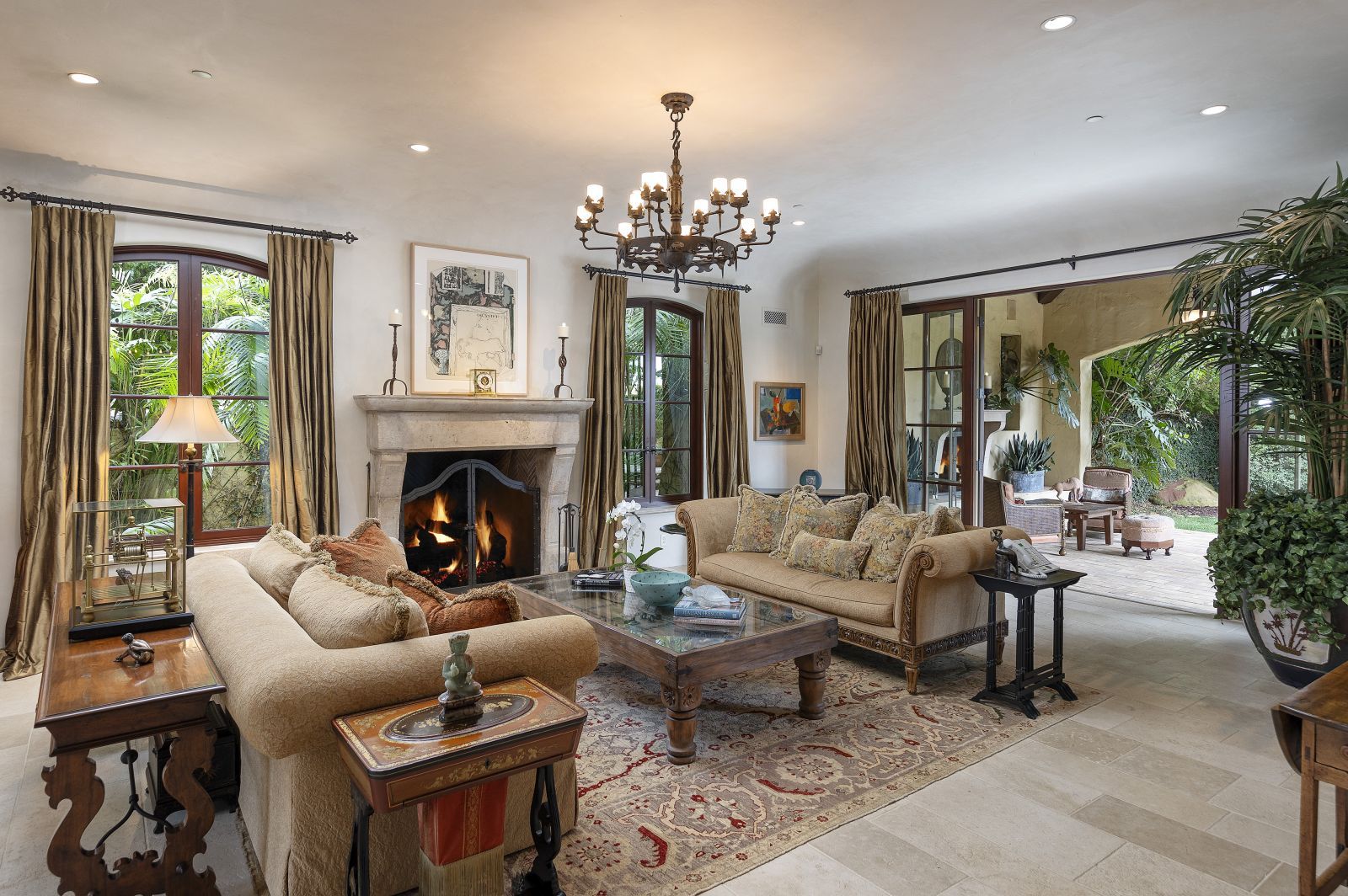 A sunken living room inside a luxury Montecito estate with a fireplace, a chandelier, comfy furniture and sliding glass doors that lead to a patio.